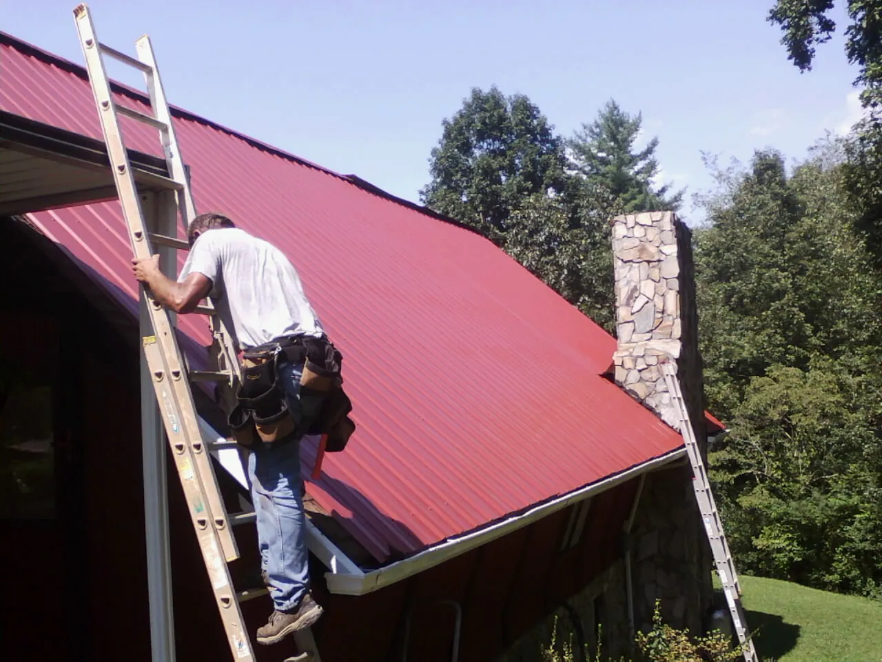 Roofer on a ladder beside a red metal roof and stone chimney — Soffit & Fascia Repair by Roofing Brothers in Amelia